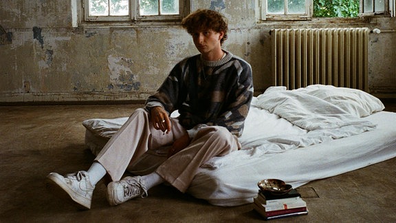Musiker Francis sitzt in einem heruntergekommenen Raum auf einer Matratze mit Zigaretten und Büchern ausgestattet