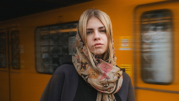 Künstlerin ELLICE hat blondes, langes Haar, das sie unter einem Schal trägt, sie steht draußen vor einer vorbeifahrenden U-Bahn 