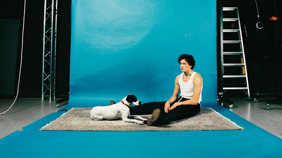 Künstler Levin Liam sitzt mit einem Hund auf einem grauen Teppich vor einer blauen Fotowand in einem Studio, er trägt eine schwarze Jeans und ein weißes Tanktop