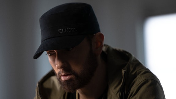 Eminem mit Mütze auf dem Kopf, Blick gesenkt und dunklen Haaren.