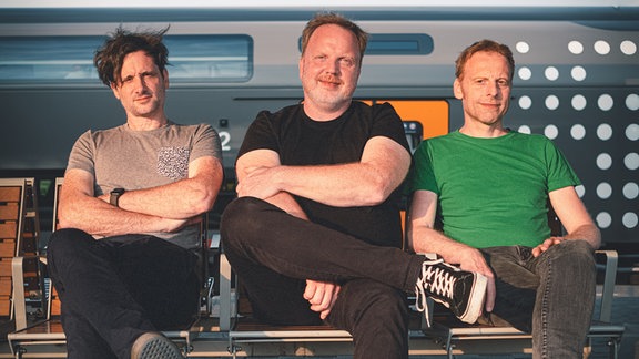 Die drei Mitglieder der Indie-Band Kapelle Petra sitzen mit überschlagenen Beinen auf einer Bank an einem Gleis vor einen Zug