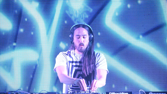Steve Aoki steht auf einer Bühne, trägt Kopfhörer und arbeitet gebannt an seinem DJ-Pult.