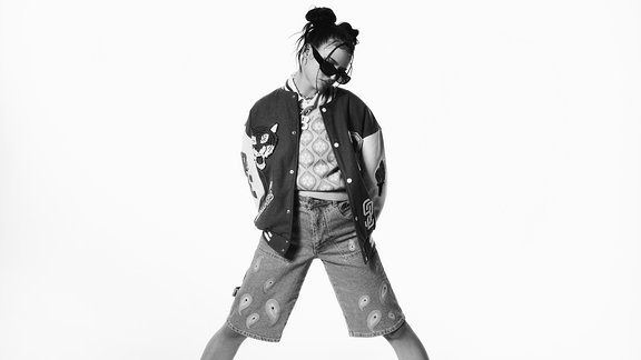 Musikerin Nina Chuba steht breitbeinig, mit Händen in den Taschen und mit Sonnenbrille auf da, schwarz-weiß Bild