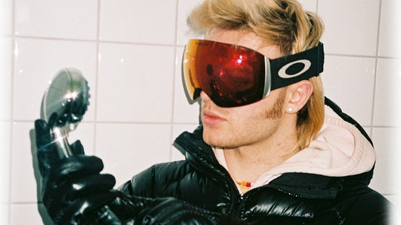 Sänger Ski Aggu mit Ski-Brille, dicker schwarzer Daunenjacke und einem alten Telefonhörer in der Hand