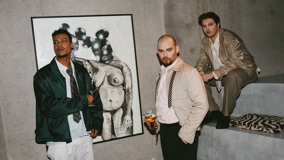 Die drei Bandmitglieder von "Schorl3" stehen in einem grauen Raum, hinter ihnen hängt ein Kunstwerk.