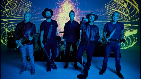 Band Pearl Jam (fünf Mitglieder) in blauem Licht, stehen frontal zur Kamera nebeneinander mit ihren Instrumenten vor gelber feinliniger Lichtinstallation 